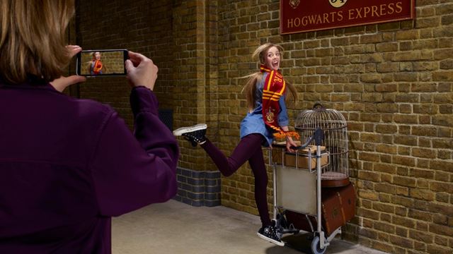 Une dame s'accrochant au chariot de bagages du quai 9¾, portant une écharpe Harry Potter, sous le panneau "Poudlard Express".