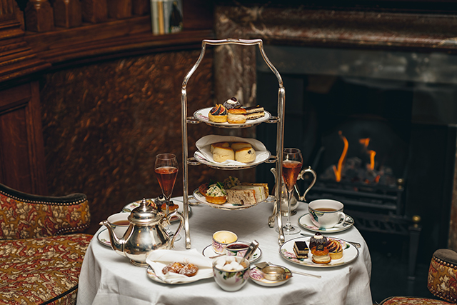 A three-tiered cake stand full of cakes and sandwiches, surrounded by plates, cups and saucers, on a table with a white table cloth, in front of a lit fire in an open fireplace.