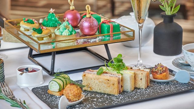 Trays of green christmas tree desserts, pink pudding baubles and finger sandwiches sit next to a glass of champagne.