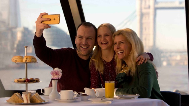 A family of three poses for a selfie while on a river cruise, with a three-tiered cake stand featuring sandwiches, cakes and scones in the foreground, and the river Thames and Tower Bridge in the background.
