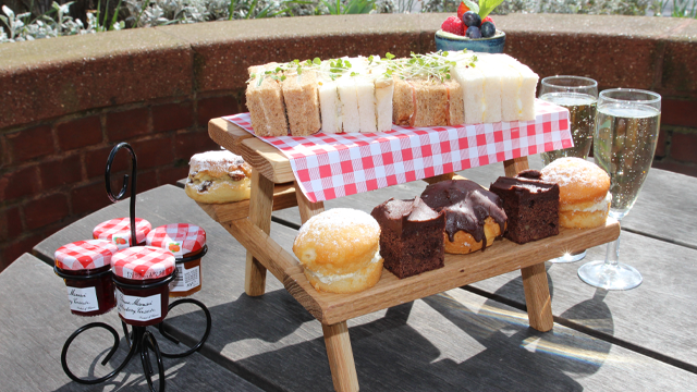 Cakes and sandwiches displayed on a mini pincic bench covered in a small tablecloth.