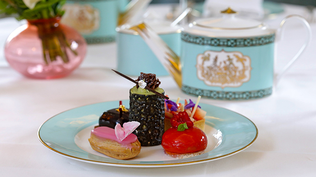 A small selection of colourful, dainty cakes on a plate with a turquoise rim, with a teapot of the same colour in the backround.