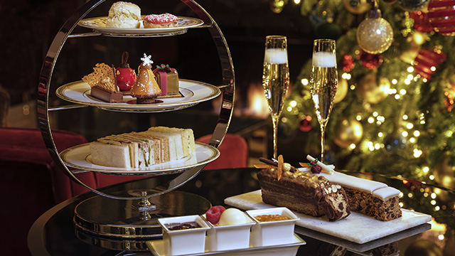 A three-tiered platter with sandwiches and Christmas-themed cakes, with Christmas cake and glasses of sparkling wine to the side, and a Christmas tree decorated with lights in the background.