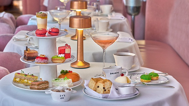 Festive-themed cakes and sandwiches on a three-tiered white cake stand plus a cocktail, scones and cups and saucers alongside on a table. The iconic pink seats of The Gallery at Sketch in the background.