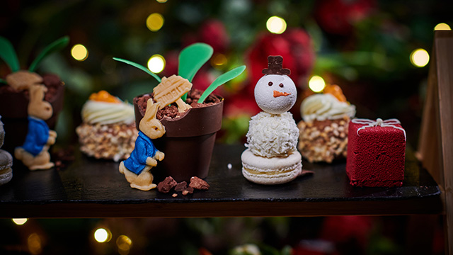 Small cakes, including some shaped in the form of Peter Rabbit, a snowman and a cube, on a wooden platter, with Christmas lights in the background.