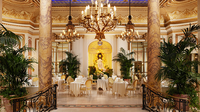 A view of the Palm Court, including palm trees on either side of marble columns, a marble floor and dining tables with white tablecloths.