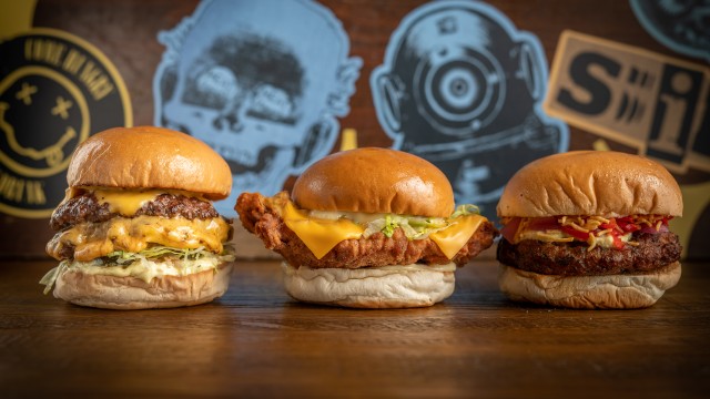 Three burgers laid out on wooden table with blue and yellow stickers on the wall in the background.