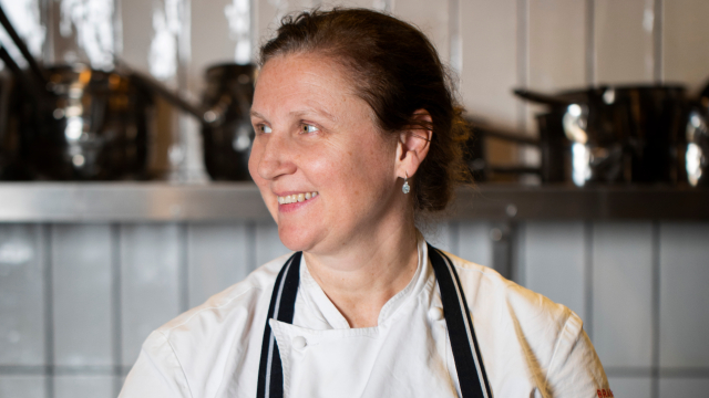 Angela Hartnett is posing for the camera in a tiled kitchen, with pan and pots on shelves in the background.