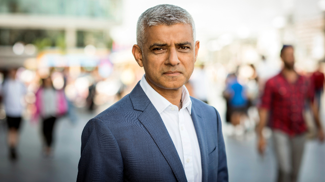 Sadiq Khan is facing the camera, wearing a suit and white shirt, with passer-by blurred in the background.