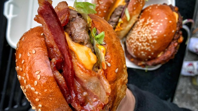 Close-up of a juicy cheese and bacon burger with more burgers in a white take-out box in the background.