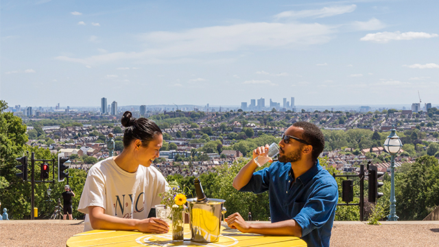 Two people with drinks sit on a yellow picnic-style table on a sunny day, with London's skyline behind.