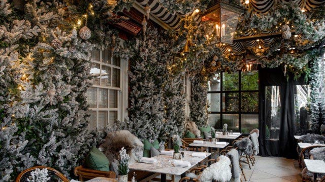Snow-frosted pine treed line a terrace of tables and chairs, which have fur throws draped over them.