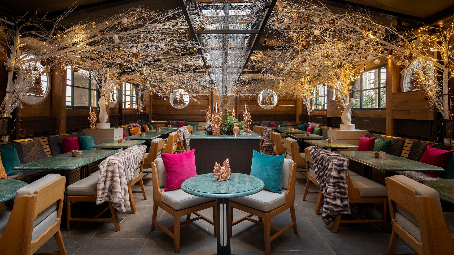 A wide-angle view of the inside of The Elyx Chalet, with a table and two chairs in the centre and illuminated tree branches with baubles lining the wooden walls.