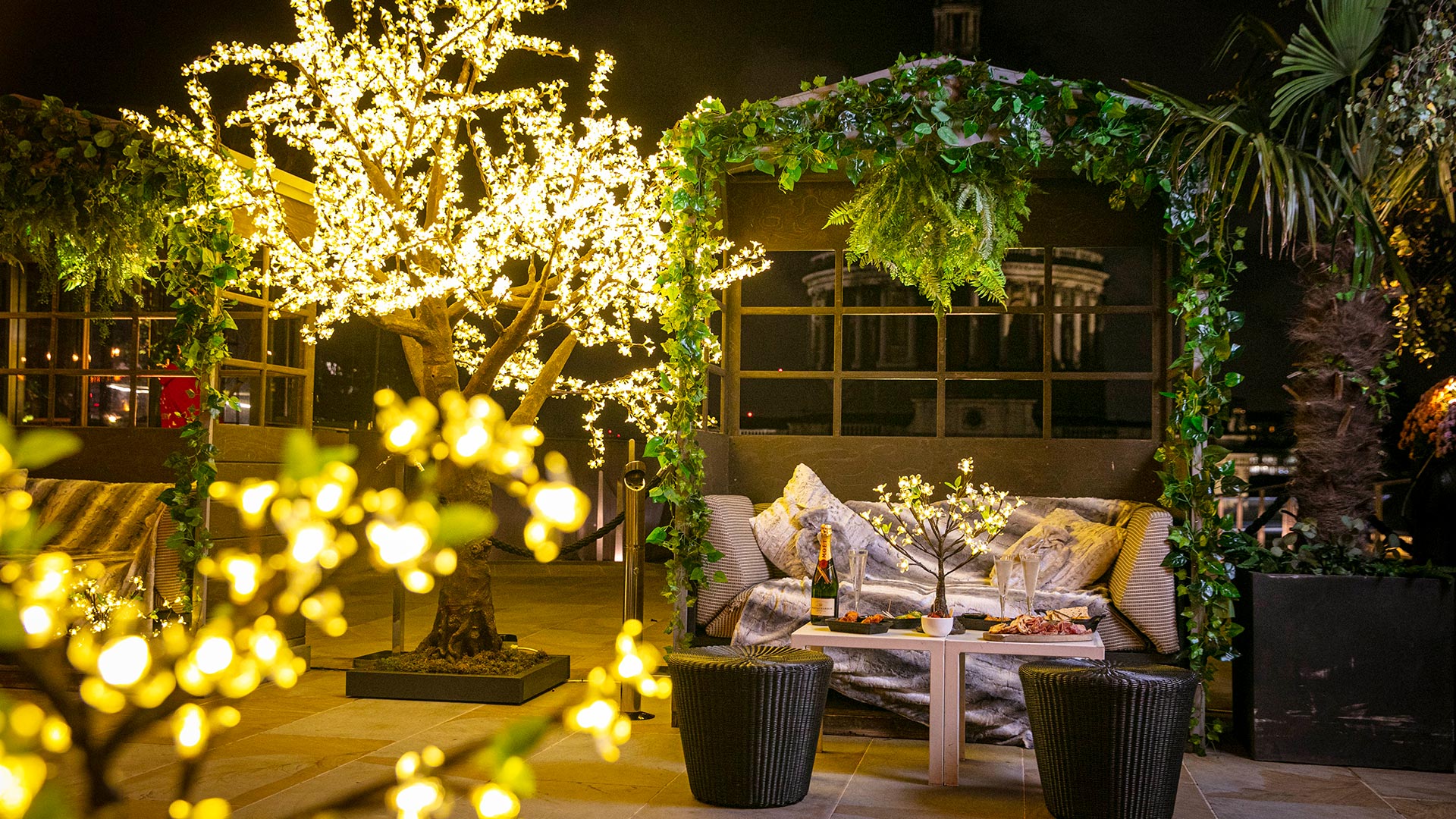 A view of Madison's Enchanted Woods winter terrace, with illuminated fairy lights around trees and a table with a bottle and glasses of champagne.