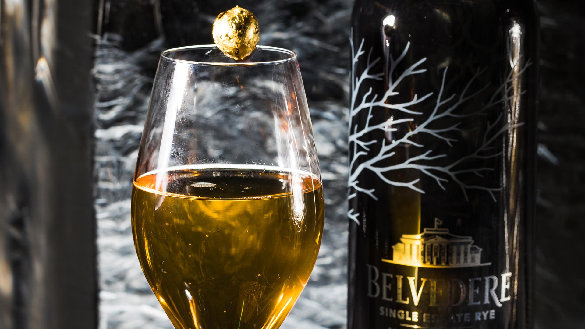 A cocktail glass is filled with the yellowish Belevedere + Ice cocktail, with a gold leaf ball balanced on top, and a bottle of Belvedere vodka in the background.