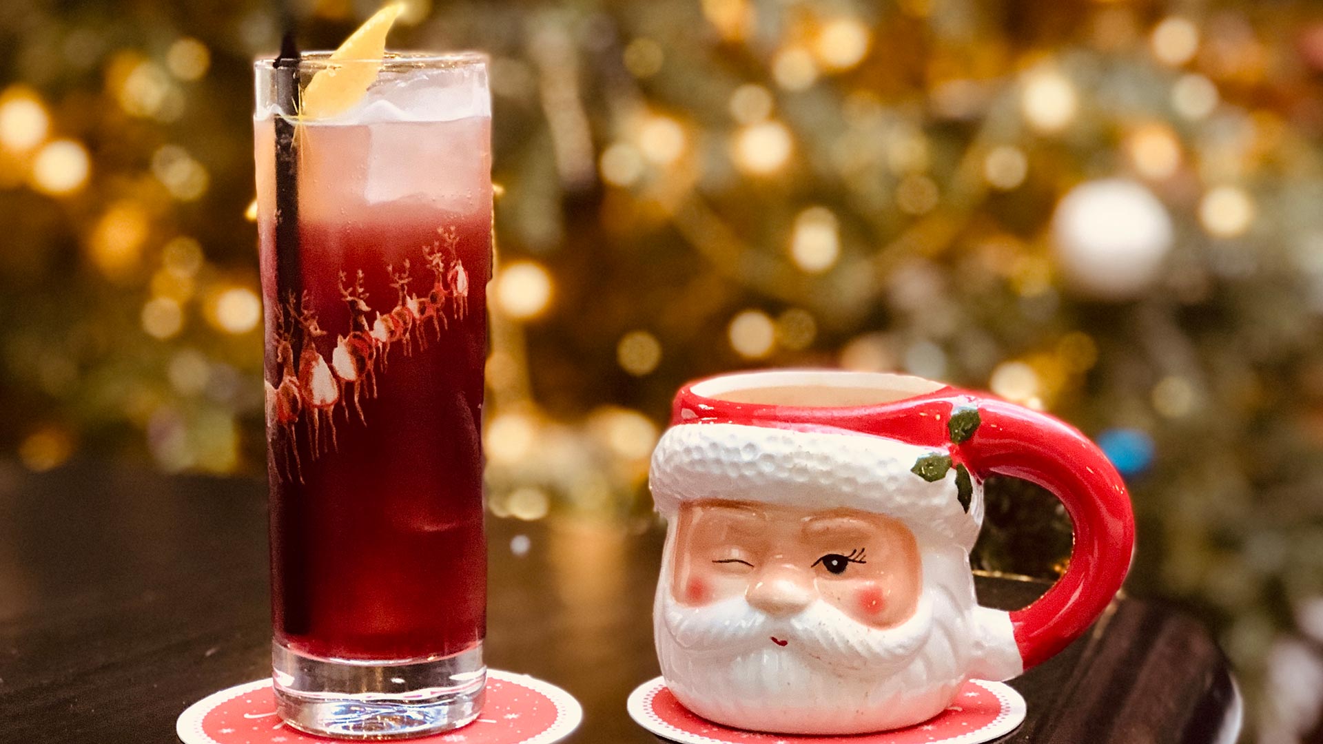 A ruby-red-coloured cocktail topped with a slice of lemon stands next to mug with a winking Santa face.