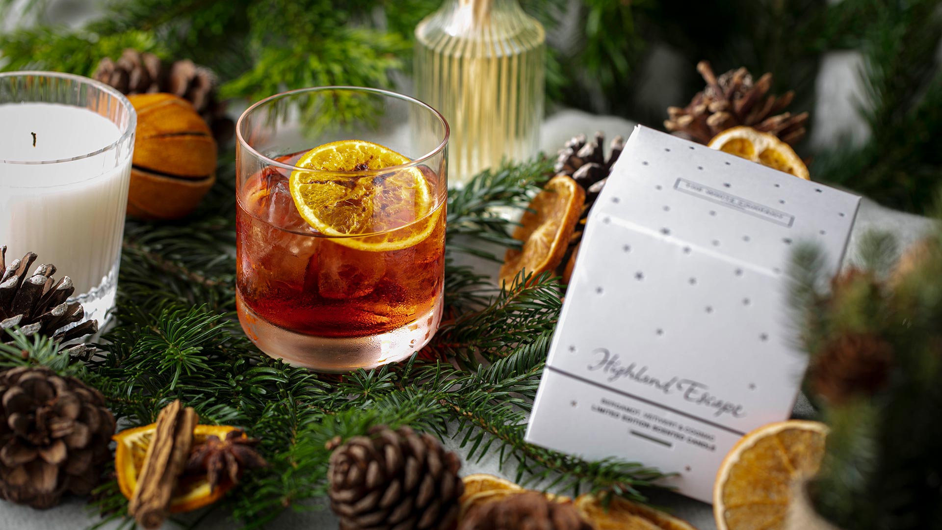 A red cocktail in a tumbler with a slide of dried lemon sits on some fir leaves, surrounded by candles and pine cones.