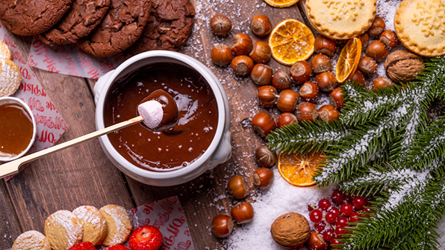 An aerial view of festive food laid out on a table, including a marshmallow being dipped in chocolate fondue, hazelnuts, cookies, mince pies and dried orange slices.