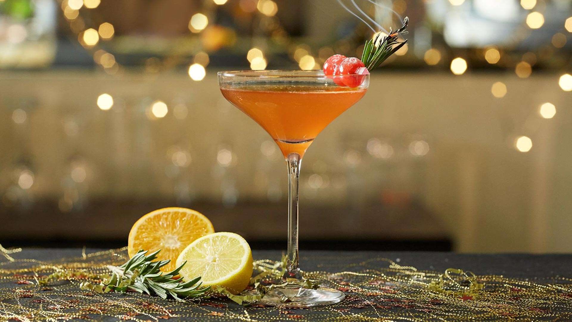 An orange-coloured Christmas Kiss cocktail, featuring cranberries and a smoking sprig of rosemary as garnish.