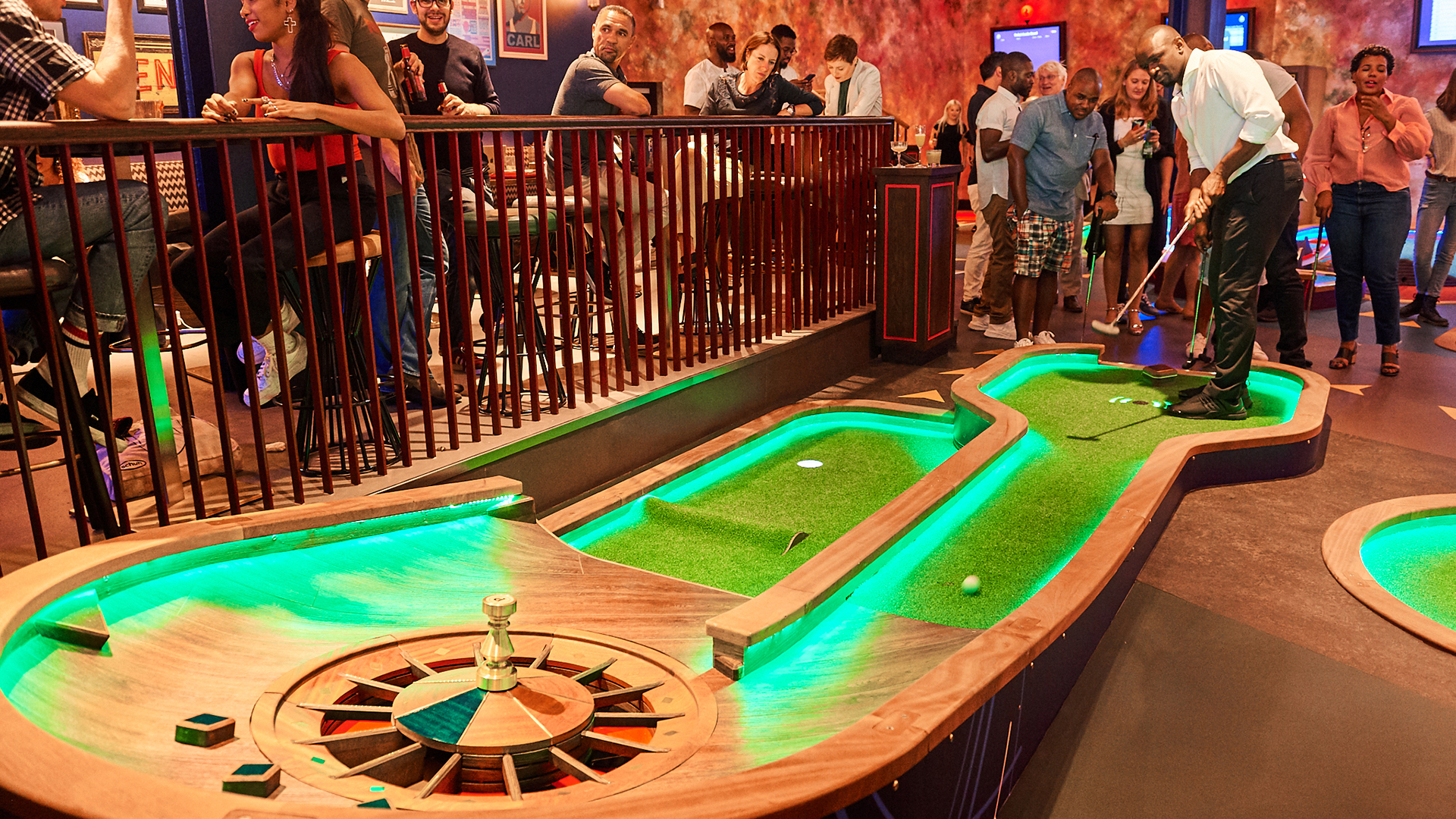 A man takes a putt on one of Puttshack's brightly lit mini golf holes, while people look on.