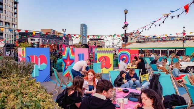 People enjoy drinks on the brightly coloured tables of East rooftop bar on a sunny day in London.