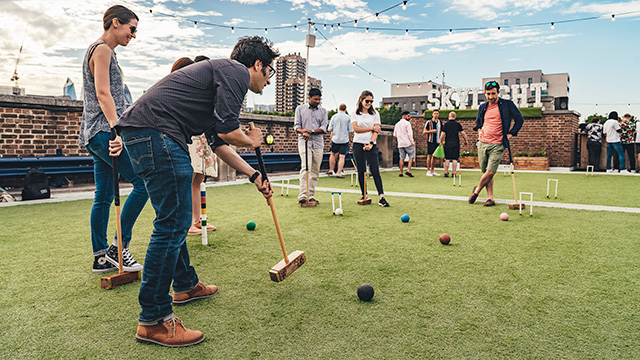 A person takes a shot using a mallet, while being watched by friends, during a game of croquet at Skylight Tobacco Dock in summer.