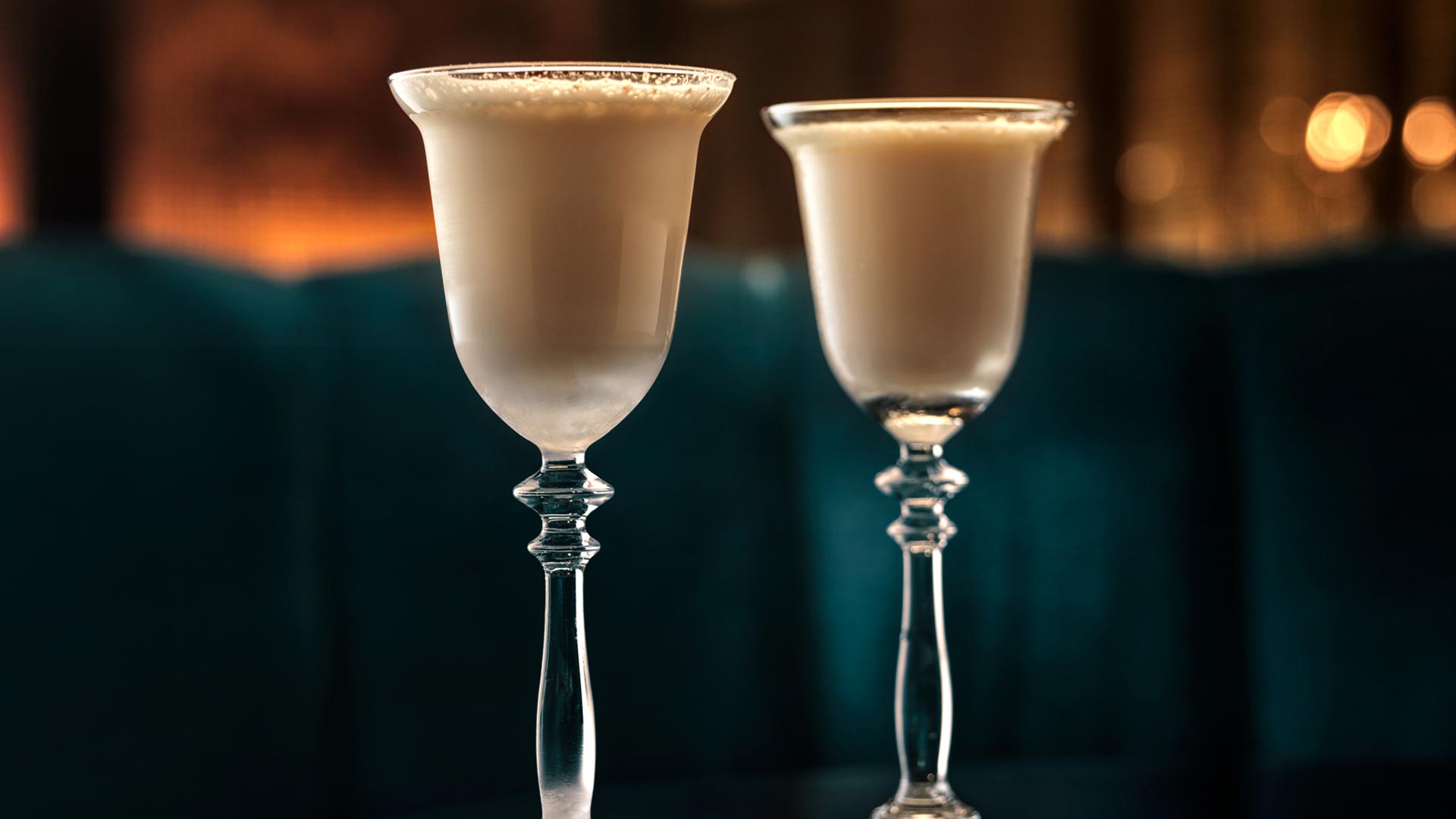 Two tall cocktail glasses, filled with the light-brown-coloured St James Bar's Milky Cat cocktail, against a dark background with lights.