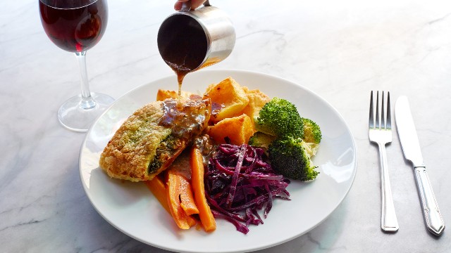 A plant-based roast with vegetables on a white plate, with gravy being poured on top of it and a glass a red wine and silver cutlery in the background.