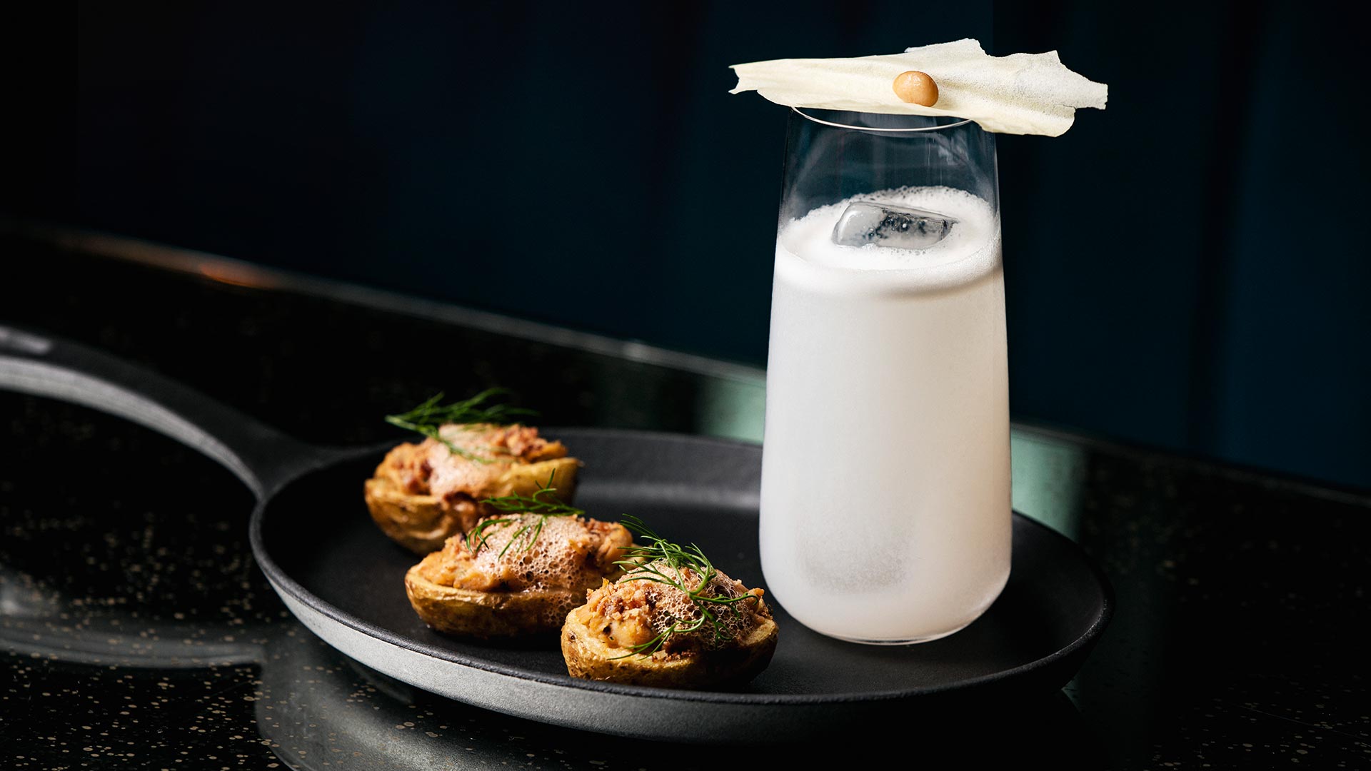 A white-coloured cocktail with a balanced garnish on top, with three small canapes by the side, all presented on a frying pan.