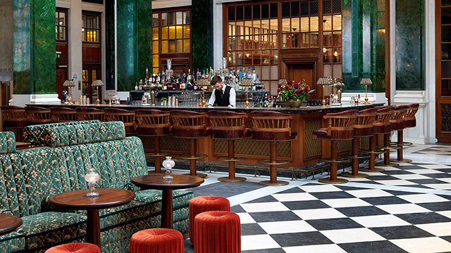 The American-style Nickel Bar at The Ned with elegant green sofas and interior and a bartender serving drinks