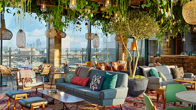 Das Interieur der Rooftop-Bar The Nest im Treehouse London Hotel mit Möbeln im Boho-Stil, Teppichen, Hängeleuchten und von der Decke hängenden Pflanzen sowie der Skyline von London im Hintergrund.