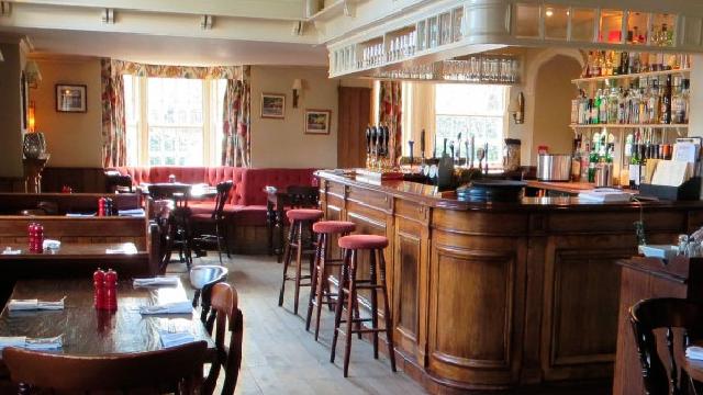 The bar at The Plough, with a traditional wood bar and stools with red fabric on the right, and pub dining tables on the left.