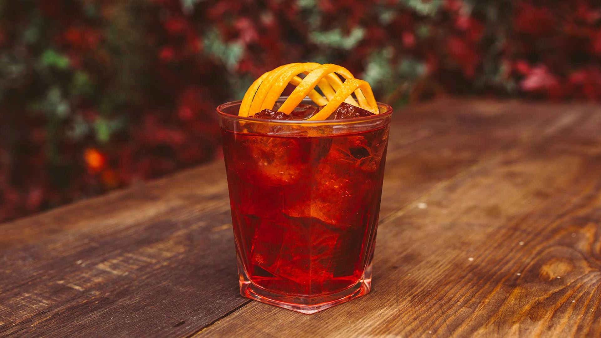 A red cocktail in a tumbler glass with decorative orange peel on top, sits on a table, with a Christmas tree in the background.