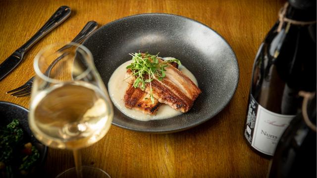 A table with a black plate with a fine dining dish of pork and sauce next to a bottle of wine and glass of white wine.