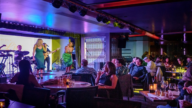Indoor bar with two female singers and a live band on stage, with people sat on small tables watching the stage