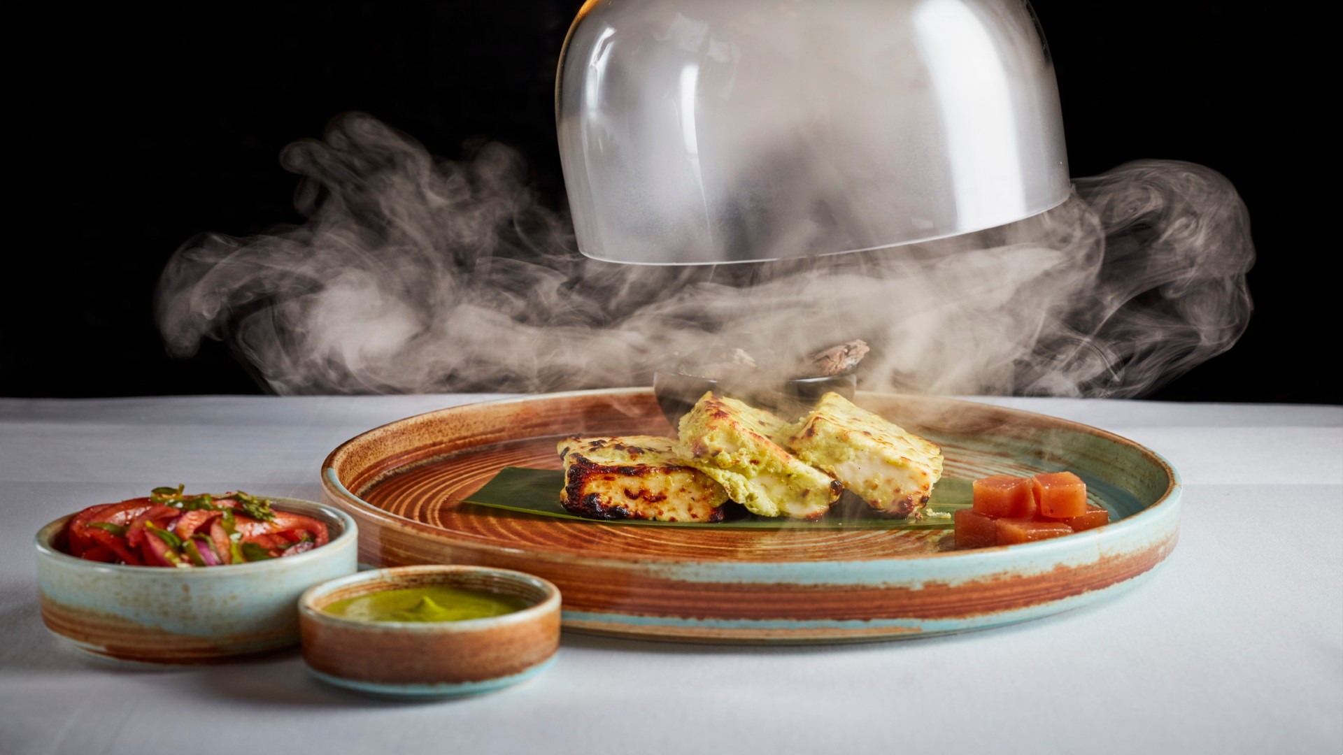 A cover is being lifted from a plate of food and steam billows out across the table.