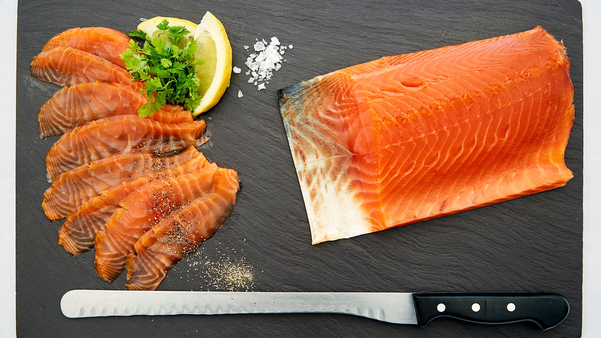 A pink tranche of smoked salmon lays on a slate platter, with slices of smoked salmon and lemon alongside.