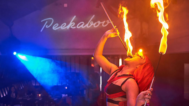 A woman on stage, a fire-breather, is holding two wooden sticks with a flame at a top while fire comes out of her mouth. 