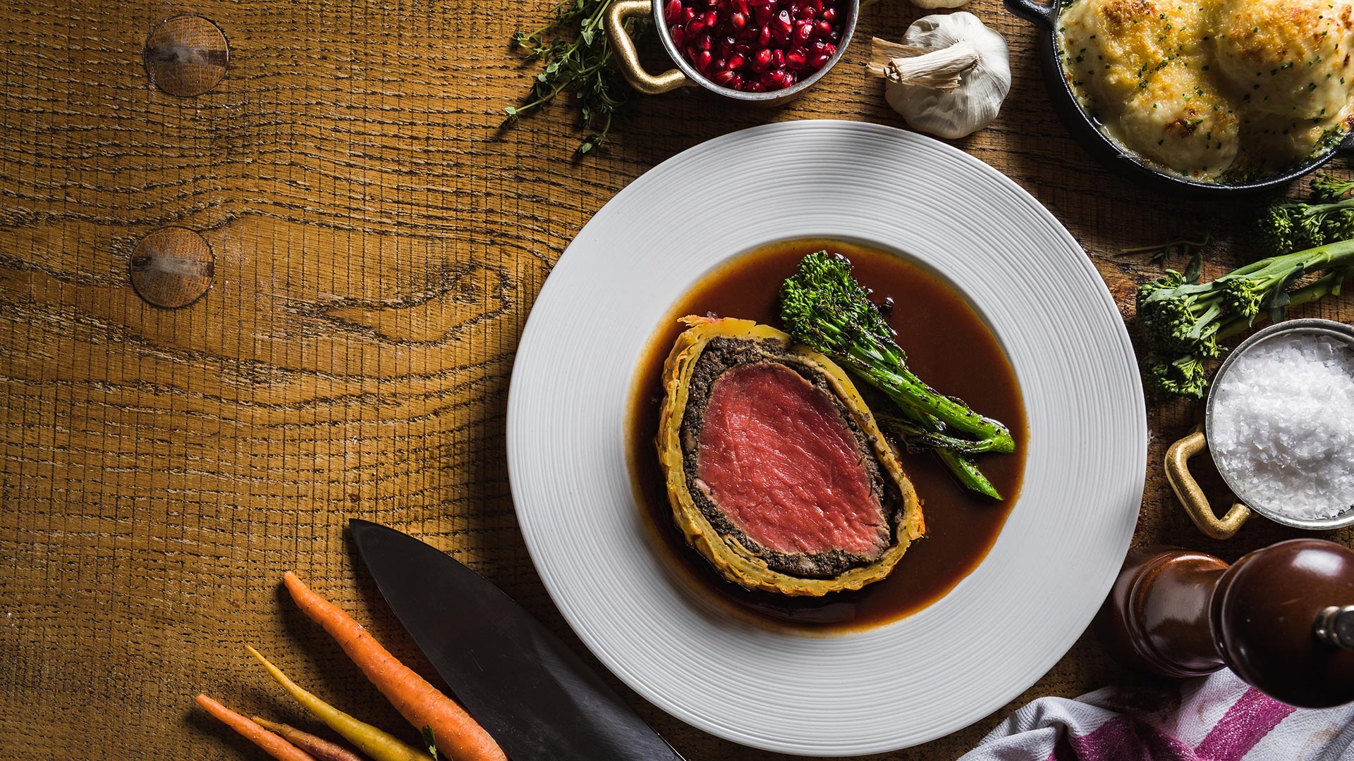 The Kitchens' beef Wellington, served rare, and long-stemmed broccoli sit on a plate with a dark red wine reduction sauce. The plate rests on a wooden surface surrounded by raw ingredients such as carrots, pomegranate and carrots.