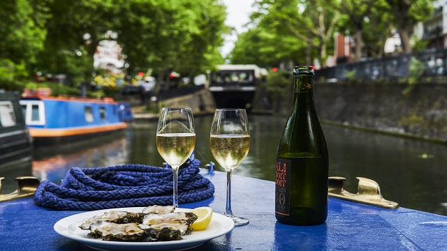 A landscape view of a seafood platter and bottle of wine on a barge, with the canal in the background blurred