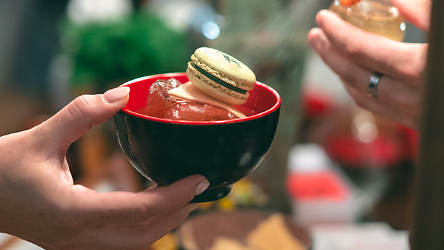 A hand holds a black and red Chinese rice bowl, filled with ice and a macaron.