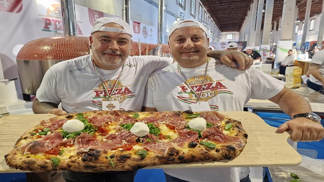 Two men in waiter uniform, holding a board with a metre long pizza on
