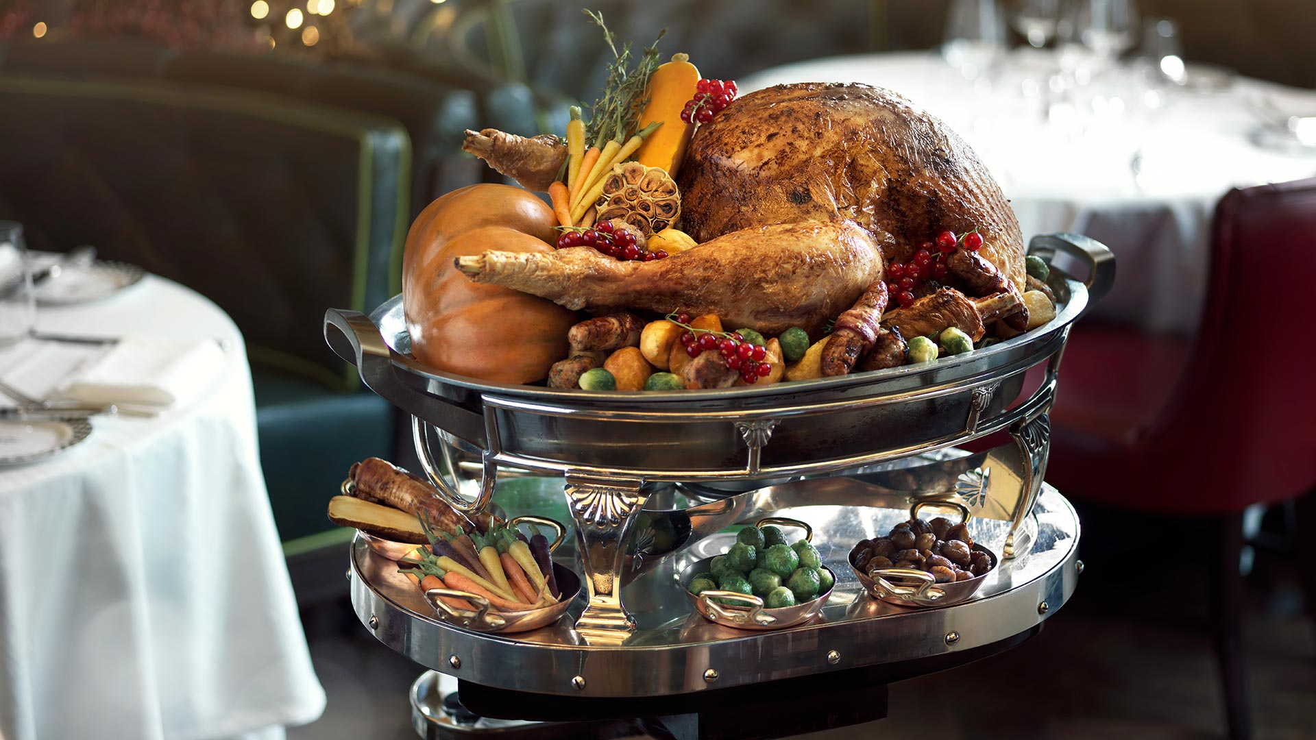 A golden roast turkey sits on the top tier of a two-tiered silver platter, surrounded by garlic, redcurrants, pigs in blankets and vegetables, at Simpson's in the Strand. The bottom tier includes brussels sprouts and chestnuts.