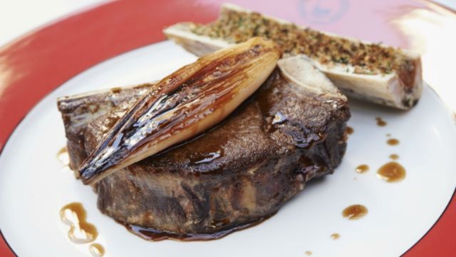 A tender steak on a white plate with a grilled onion and stuffed bone marrow with a light jus dotted around the plate.