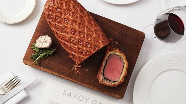 A white table with white plates and a brown chopping board with a brown and pink beef wellington.