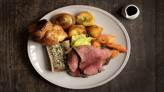 Roast beef served on a white dinner plate, with roast potatoes and vegetables, and a pot small jug of gravy