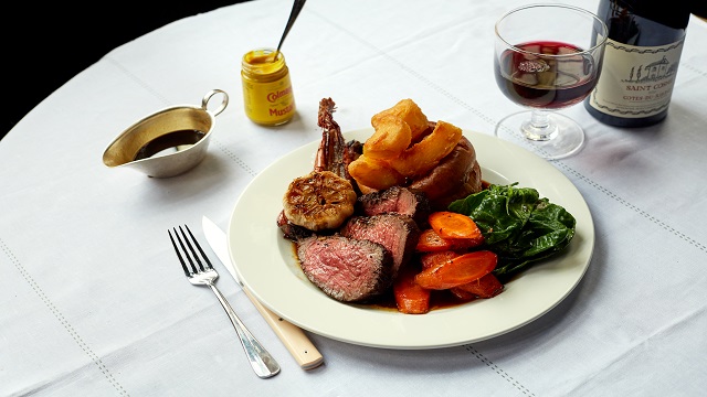 A clean white table cloth with a beef roast dinner plated up, surrounded by a glass of white wine, gravy boat and mustard pot