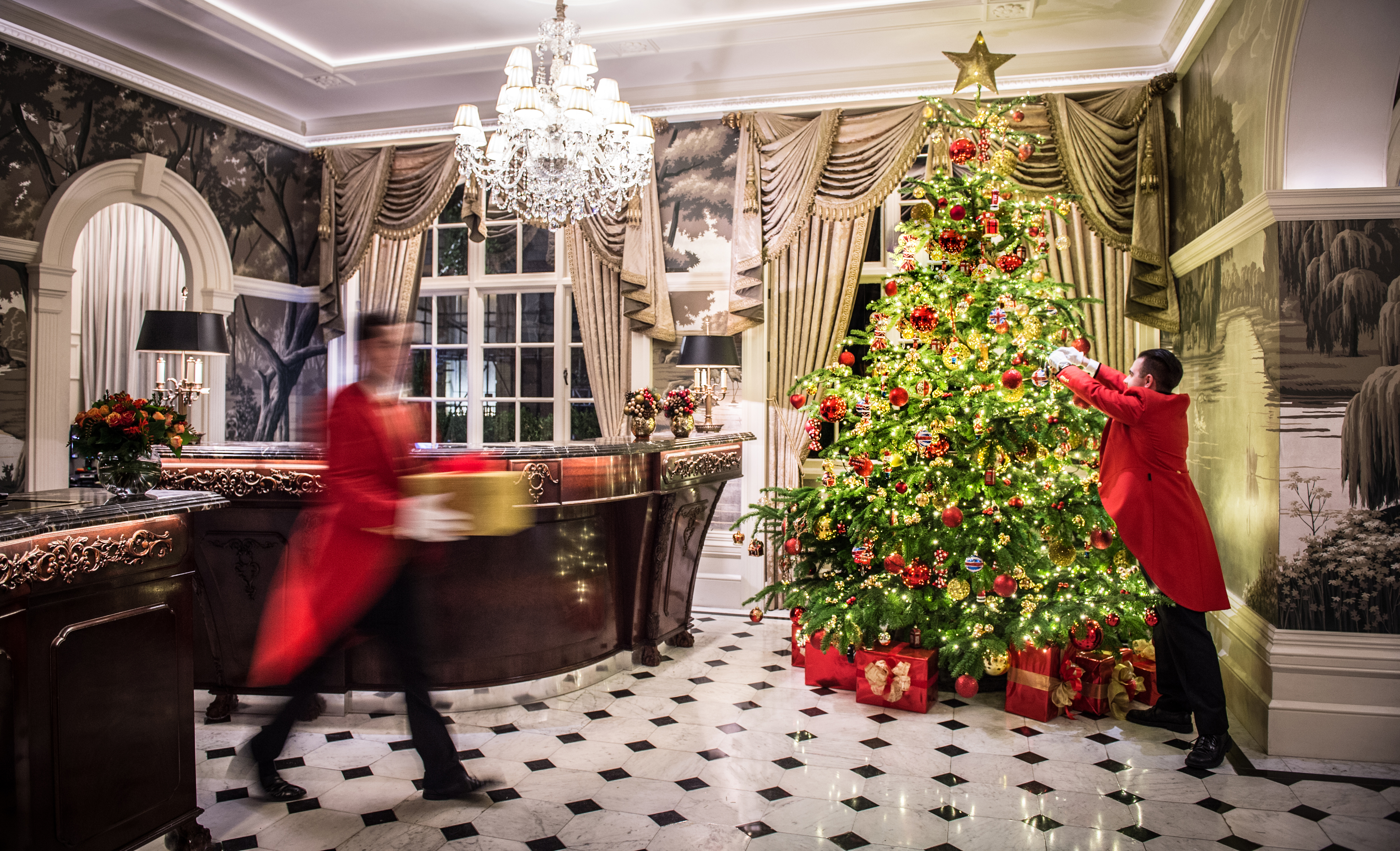 A butler, dressed in a red coat and black trousers, decorates a Christmas tree in the lobby at The Goring, featuring a chandelier and black and white-tiled floor. Another butler walks past, carrying a present.