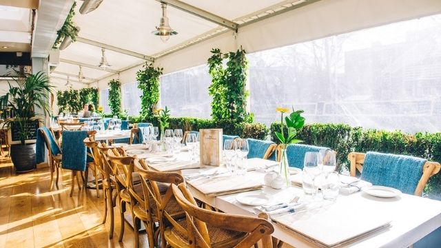 Tables and green foliage line a sunny outdoor dining space at London restaurant The Summerhouse.