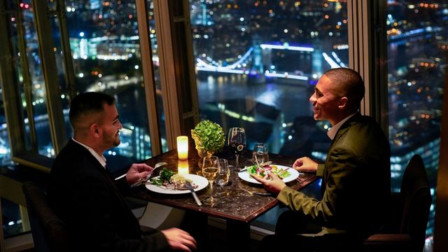 Two men sit at a table enjoying a dinner whilst overlooking london bridge at night.